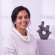 Smiling South Asian woman in her late 30s, headshot portrait with a soft lavender and white background.